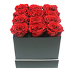 This image depicts a striking, minimalist arrangement of twelve deep red roses presented in a square black gift box, captured against a bright white background that makes the colours and shapes stand out in sharp relief. The roses, likely preserved for long-lasting beauty, are arranged in an exact grid of three by four, each head touching the next so that the viewer sees a continuous field of crimson blooms. Every rose is in perfect full bloom, with densely packed, velvety petals spiralling outwards from a darker centre to gently lighter outer edges, creating a rich texture you can almost feel with your fingertips. They sit slightly above the rim of the box, suggesting skilled florist placement to achieve a uniform, level top while still allowing the curves of each bloom to be clearly defined. The box itself is a matte black square with crisp edges, its surface smooth and unembellished, conveying understated, modern luxury. Near the lower edge of the container runs a slim white stripe, wrapping all the way around like a precise graphic accent that echoes the clean lines of contemporary interiors found in and around Golders Green. Soft, even lighting falls across the arrangement, casting only the faintest shadows beneath the petals and along the sides of the box, which enhances depth without introducing harsh contrast. The result is an image that feels calm, composed, and sophisticated, as if capturing the moment before this elegant rose box is delivered to a doorstep off Finchley Road or set on a dining table for an intimate anniversary dinner. It speaks of romance, gratitude, and cherished connections, all distilled into twelve flawless red blooms.