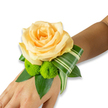 A tender wrist corsage rests along the inner wrist, the centerpiece a buttery yellow rose unfurling in luminous, sunrise-like layers; its velvety petals catch soft light and suggest the gentle warmth of a wedding morning. Nestled beneath the rose are two lively lime-green button chrysanthemums, their compact, pom-pom forms offering a cheerful textural counterpoint to the smooth rose face. Glossy, dark green leaves - the sturdy, salal- or ruscus-like foliage - cradle the composition, giving it depth and a fresh, grounded silhouette. A striped, variegated leaf is artfully curled in a ribboned arc to one side, its cream-flecked stripes and architectural fold adding a modern flourish that increases the corsage's visual movement. The flowers appear freshly cut; you can almost sense the faint scent of rose and the green, grassy hint that follows when foliage is handled. The arrangement sits against a clean white background so the colours read clear and true, and while the fastening is not visible, the design suggests a discreet elastic or ribbon beneath the leaves. This small, handcrafted floral wristlet radiates an intimate, anticipatory mood - ideal for first dances, vows, or an evening celebration where subtle elegance is desired. As a local touch, this style is often chosen by brides stepping out near Golders Hill Park or guests arriving by Golders Green station, a favourite for quiet, refined moments in the neighbourhood. The overall effect is soft yet striking: equal parts handcrafted embrace and wearable art that honours both the occasion and the living flowers.