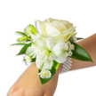 A delicate wrist corsage sits against a clean white backdrop, framed on the fair-skinned wrist of the wearer as the arm extends from the lower right into the centre of the frame. At the heart of the arrangement a velvety, creamy ivory rose opens in soft spirals, its petals catching a cool, diffuse light and lending the piece a gentle, romantic glow. Just below and a little to the left a large trumpet-shaped white bloom - with the slender, slightly ribbed petal texture and pale lime-green throat of alstroemeria - provides a graceful, open silhouette that balances the rose. Small clustered buds of green promise new blooms, while airy sprays of tiny white gypsophila float like whispering clouds around the main flowers. Near the base, petite daisy-like white florets with sunny yellow centres add a sweet, vintage note and a point of bright contrast against the white petals.

Lush, broad deep-green leaves with a waxy sheen frame the composition, their glossy surfaces setting off the pristine whites and suggesting a freshly clipped, garden-picked quality. The blooms are secured by a chic multi-strand pearl bracelet; its round, lustrous beads wrap gently around the wrist, offering a tactile, cool-smooth band that anchors the floral design. The overall effect is one of timeless elegance - a corsage that reads as both classic and intimate, suited to weddings, proms, and formal dances. Imagine the faint, fresh scent of roses and summer stems, and the quiet pride of slipping this onto your wrist before an evening in Golders Green or a photograph beneath the soft light of a nearby park.