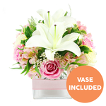 A carefully composed bouquet of white and pink flowers sits in a clear square glass vase, photographed against a bright white background that brings every detail into sharp focus. At its heart are striking white Oriental lilies, their large petals fully open and gently flared, each bloom showcasing prominent reddish-brown stamens that lend the arrangement a refined, modern edge. Surrounding the lilies, numerous pink roses gather in a harmonious cluster, some still in bud form with tightly wound centres, others opening into soft, layered rosettes that suggest affection and warmth, the sort of design often chosen by families in Golders Green to celebrate birthdays or express heartfelt thanks. Interwoven among the roses are graceful stems of alstroemeria in soft pink and creamy white, their inner petals delicately flecked with yellow and fine markings that reward a closer look. Small sprays of white baby's breath hover between the larger flowers like delicate lace, giving the whole design a light, cloud-like quality rather than heavy fullness. The blossoms are cradled by a rich selection of greenery: smooth, broad leaves, deeper emerald tones, and feathery fronds that add volume and a fresh garden feel, reminiscent of a calm walk through nearby Golders Hill Park. All the stems are neatly arranged in a low, square clear glass vase filled with clean water, the tidy cut ends visible and adding to the sense of freshness and professional floristry. Around the centre of the vase, a pale pink satin ribbon is wrapped cleanly and tied to form a simple, elegant band, making the arrangement appear gift-ready for same-day delivery across NW11. In the bottom right corner of the image, an orange circular badge with bold white capital lettering announces VASE INCLUDED, clearly indicating that the container is part of the floral gift.